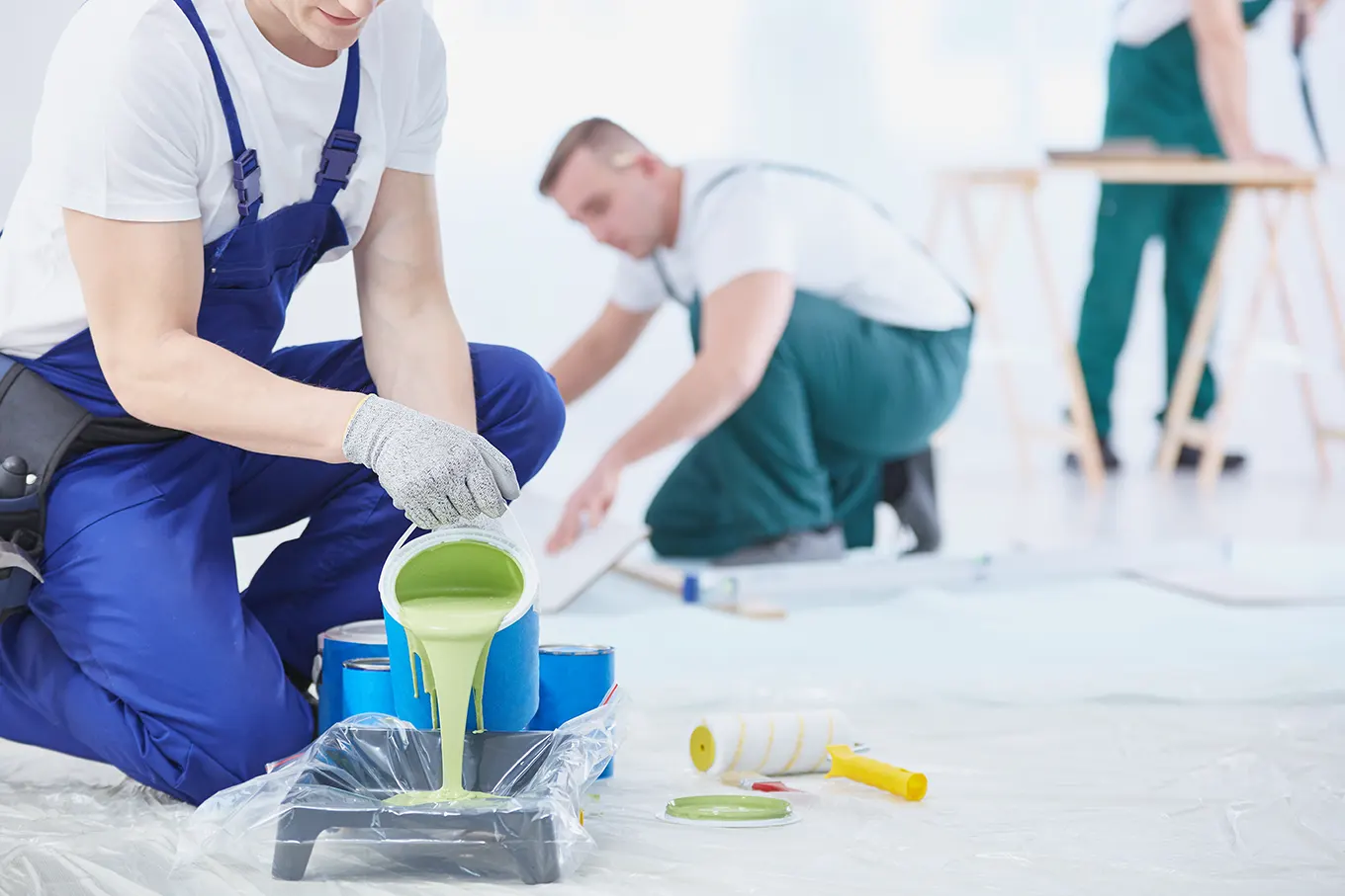 Painters in overalls pouring paint into a roller tray on a covered floor